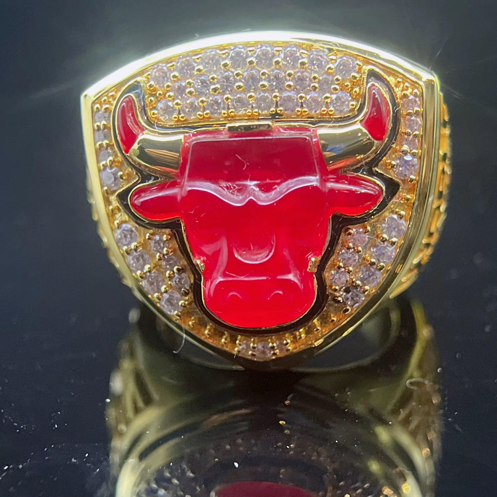Gold ring with red bull emblem and diamond accents on a reflective surface
