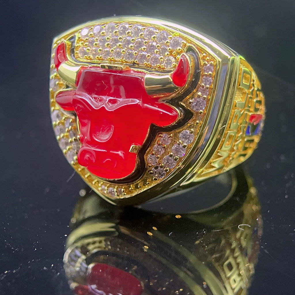 Gold ring with red bull emblem and gemstones on a reflective surface