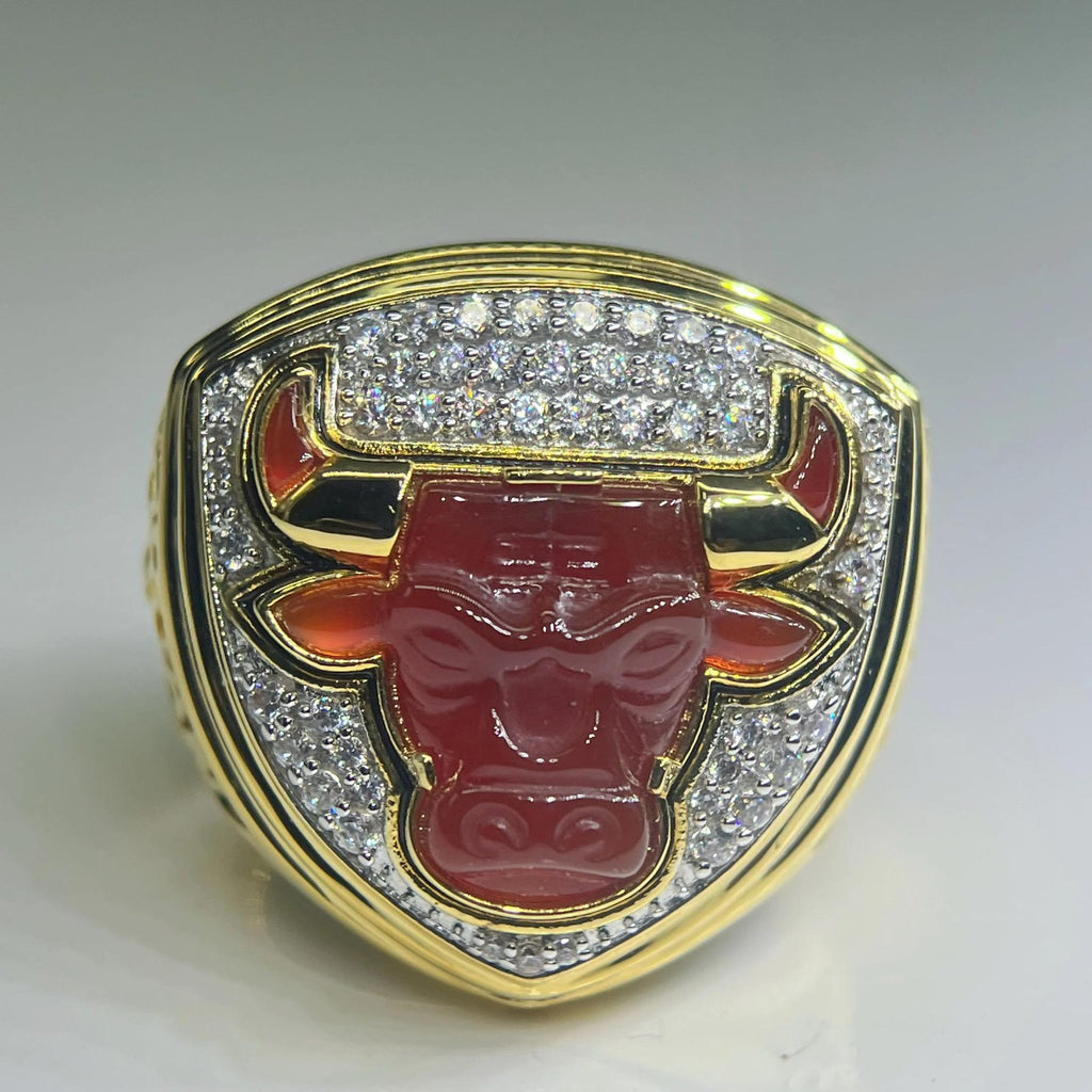 Gold ring with a red bull emblem and diamond-studded border on a white background