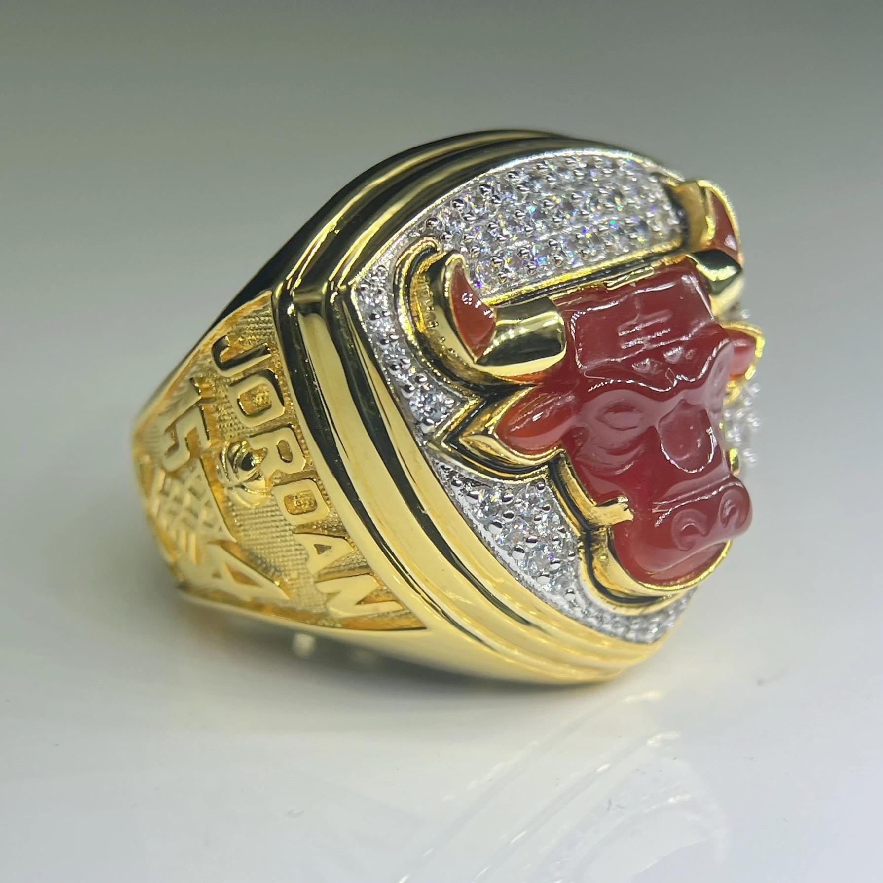 Gold ring with a red bull emblem and diamond-studded band on a light gray background