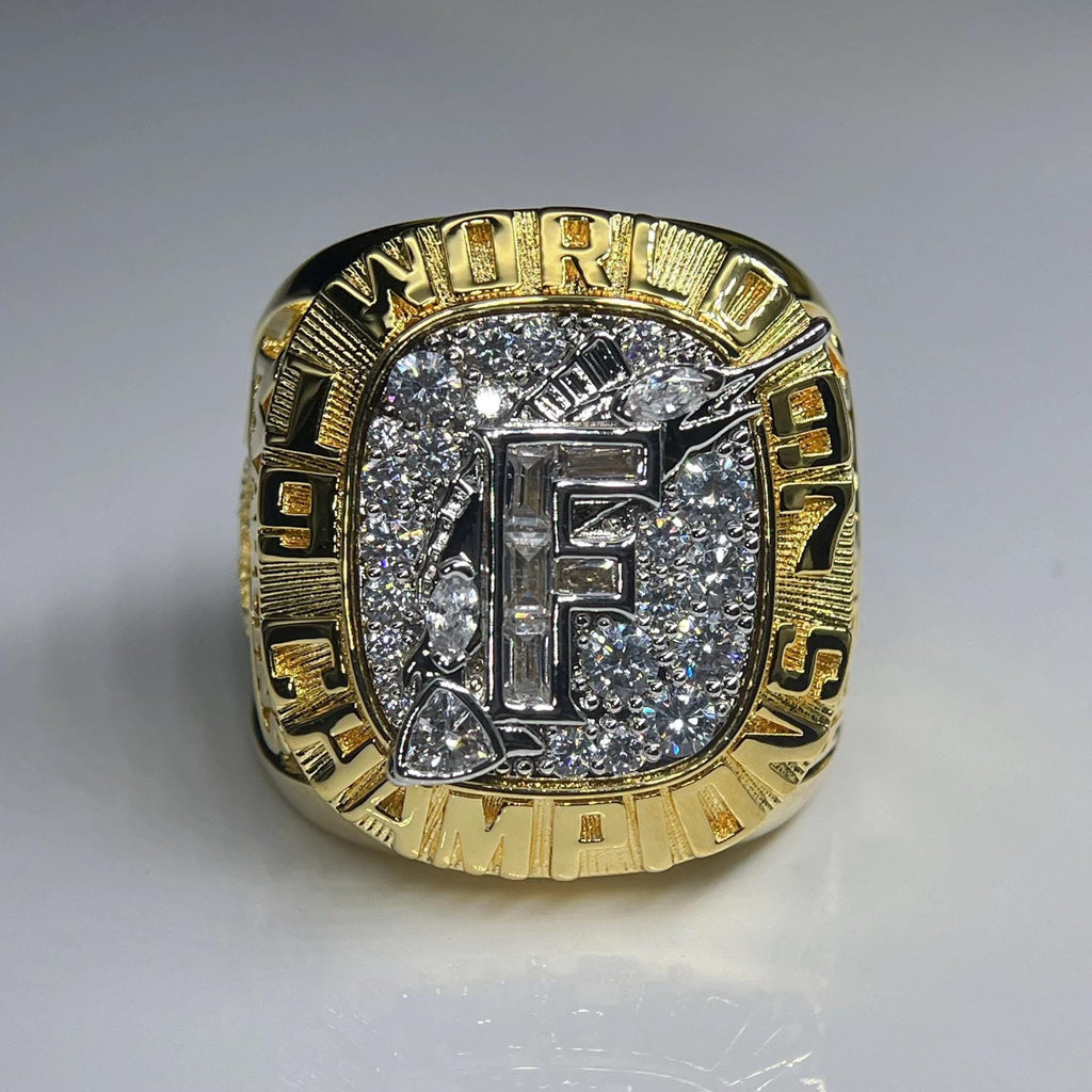 Gold championship ring with 'F' and diamond-like stones on a gray background