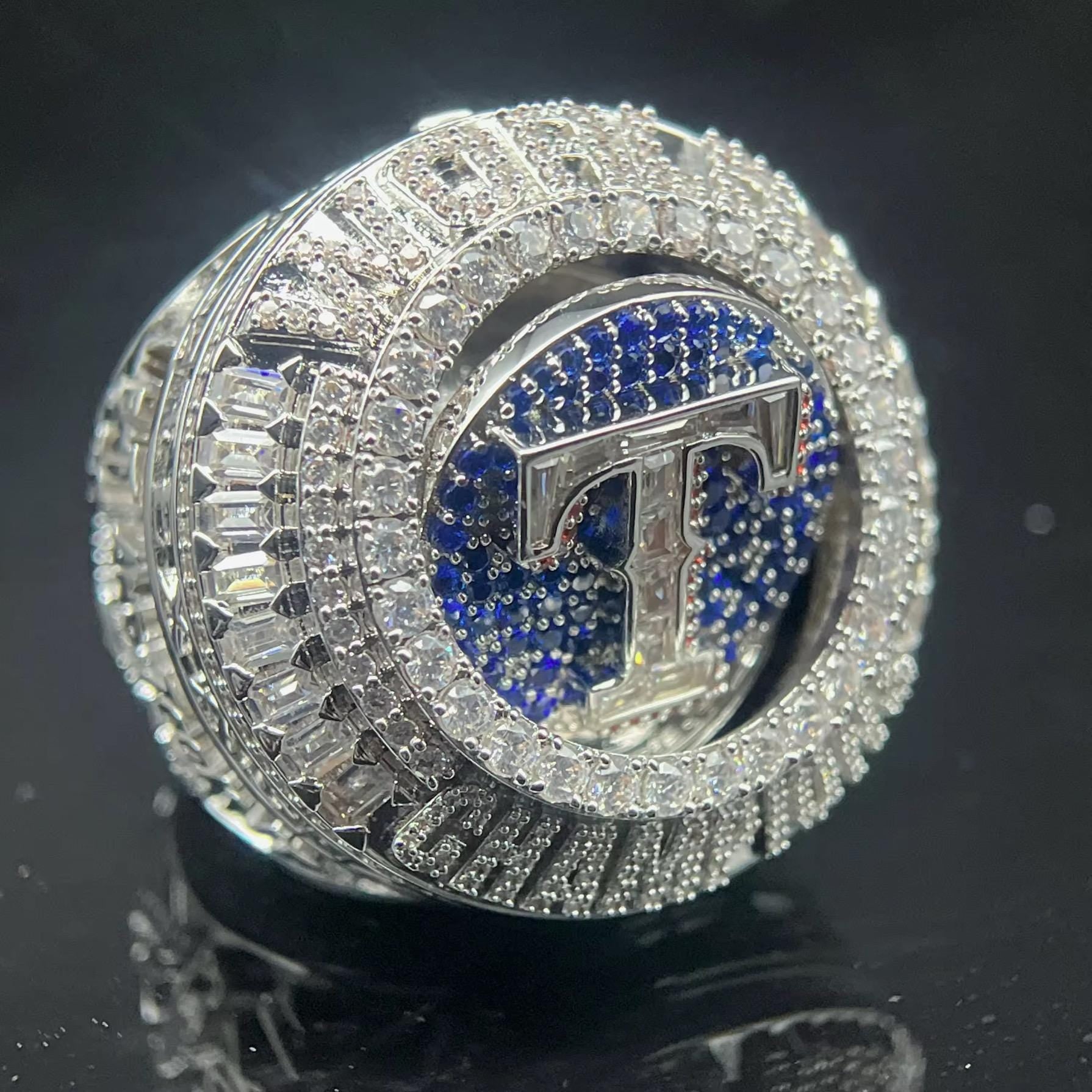 Silver ring with blue gemstone and 'T' emblem on a black background